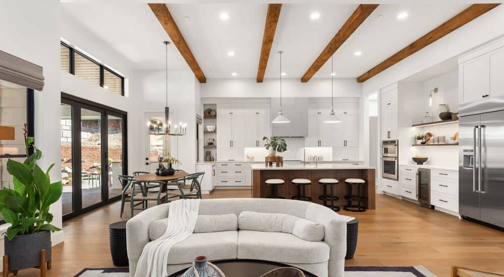 Open concept living, dining, and kitchen space featuring exposed wood beam ceilings, a dark walnut island with bar seating, white shaker cabinetry, hardwood floors, and black-framed patio doors in a Kelowna custom home.