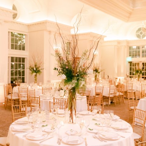 Tables adorned with white linens and tall floral arrangements are prepared for a festive evening event in a grand venue.